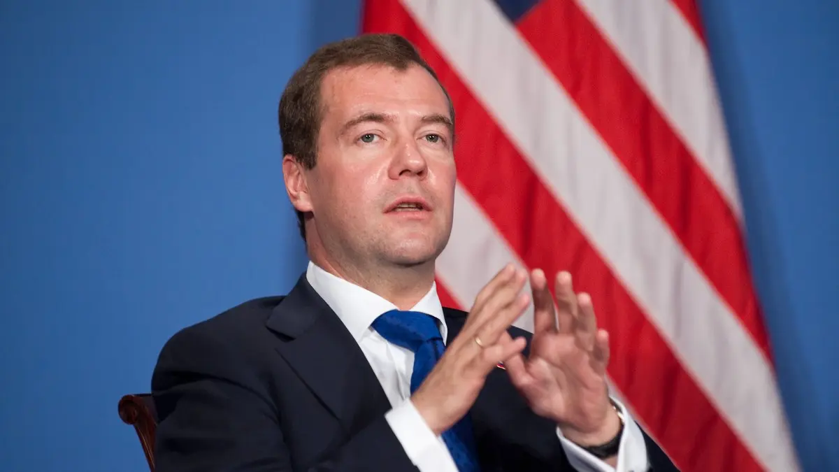 גבר לובש חליפה וידיו מורמות באמצע נאום. דמיטרי מדבדב על רקע דגל ארצות הברית