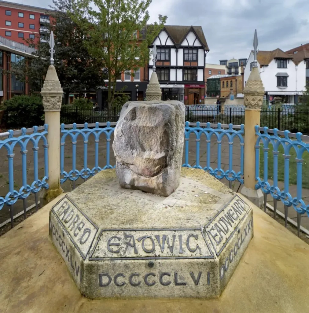 תמונה של אבן גדולה על כן במרכז שביל, מוקפת בגדר ברזל מעוטרת. אבן ההכתרה בקינגסטון שעל נהר התמזה, לונדון רבתי. צילום: © ארכיון Historic England.